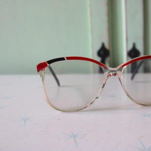 Puede incluir: Gafas de plástico transparente de ojo de gato con ribetes rojos y negros. Las gafas tienen un estilo retro.