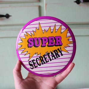 May include: A round pink and white button with a yellow starburst design. The button says "SUPER SECRETARY" in black letters.