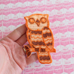 Könnte beinhalten: Ein gestickter Eulen-Patch in Orange und Braun. Die Eule sitzt auf einem Ast und hat große, runde Augen.