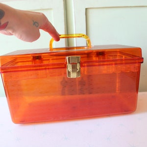 Puede incluir: Caja de almacenamiento de plástico naranja con asa amarilla y cierre dorado. La caja es transparente y tiene forma rectangular.