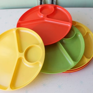 Op de afbeelding: Een stapel kleurrijke verdeelde borden in geel, groen en rood. Elk bord heeft drie compartimenten en een cirkelvormig gedeelte. De borden zijn van plastic en liggen op een wit oppervlak.