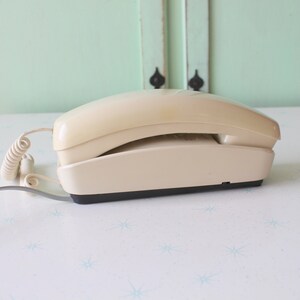 May include: A beige rotary phone with a coiled cord. The phone is on a white surface with a blue star pattern.