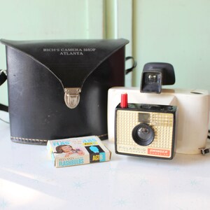 May include: A white Polaroid Swinger camera with a black leather case and a box of flashbulbs. The case has the text "Rich's Camera Shop Atlanta" printed on it.