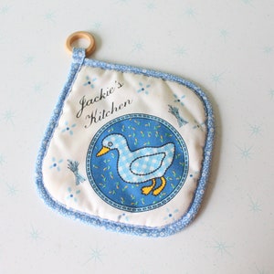 May include: A white pot holder with blue trim and a blue and white checked duck design. The pot holder has the text "Jackie's Kitchen" embroidered on it.