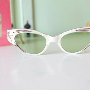 May include: White cat-eye sunglasses with a burgundy and gold design. The lenses are green.