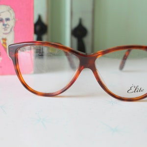 Puede incluir: Gafas marrones con montura de carey y forma de ojo de gato. Las gafas tienen lentes transparentes y la palabra "Élite" está impresa en la lente derecha. Un libro rosa con el título "Assignment: The Hideout" está en el fondo.