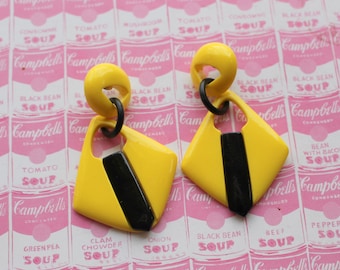 May include: A pair of yellow and black geometric earrings. The earrings are shaped like a diamond with a black stripe down the center. The earrings are hanging from a silver hook.