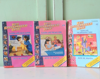 May include: Three paperback books from The Baby-Sitters Club series. The books are pink, blue, and green with white text. The titles are "Claudia and Mean Janine", "Claudia and the Bad Joke", and "Dawn and the Impossible Three".