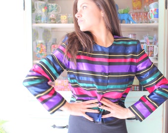 May include: A woman wearing a colorful striped jacket with a black waistband and black pants. The jacket has stripes of pink, purple, blue, green, and yellow.