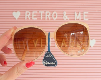 May include: A pair of peach-colored sunglasses with brown lenses. The sunglasses are being held by a hand and have a blue tag that says "Sunbrella" and "all $." The sunglasses also have the text "CR 39" on the right lens.