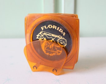 Puede incluir: Un juego de seis posavasos de plástico naranja con un diseño negro y dorado que presenta la palabra "FLORIDA" y una palmera. Los posavasos están apilados y tienen un soporte en la parte inferior.