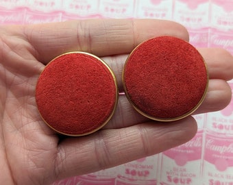 May include: A pair of red velvet earrings with gold metal settings. The earrings are round and have a slightly textured surface.