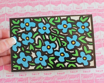 May include: A postcard with a black border featuring a pattern of blue flowers with yellow centers and green leaves on a white background.
