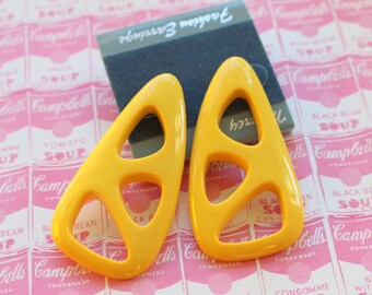 May include: A pair of yellow geometric earrings with cut-out designs. The earrings are on a pink background with a pattern of Campbell's Soup cans.