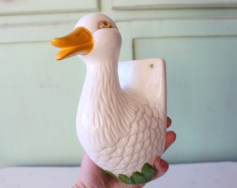 May include: White ceramic duck figurine with a yellow beak and green leaves. The duck has a hole in the back for hanging.