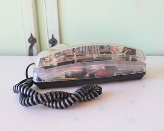 Könnte beinhalten: Ein transparentes, vintage Drehwahltelefon mit schwarzem Sockel und einem schwarzen Spiralkabel. Die inneren Komponenten des Telefons sind durch das transparente Plastik sichtbar.