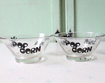 Op de afbeelding: Twee glazen kommen met een zwarte "POP CORN" opschrift. De kommen zijn leeg en staan op een wit oppervlak.