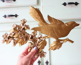 Pode incluir: Decoração de parede de metal dourado com um pássaro empoleirado em um galho com delicados acentos florais.
