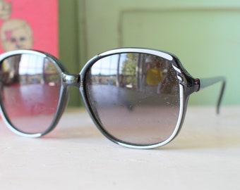 Puede incluir: Gafas de sol negras con un borde blanco alrededor de las lentes. Las lentes están tintadas de un color gris oscuro.