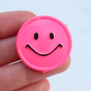 May include: A vibrant pink smiley face badge with a black smiling mouth and two black eyes. The badge is circular and has a shiny finish. A cheerful accessory.