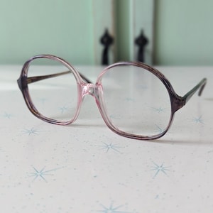 Puede incluir: Gafas vintage de gran tamaño con montura rosa y marrón. Las gafas tienen forma redonda y lentes transparentes. Las patillas son de color marrón oscuro. Las gafas están sobre una superficie blanca con diseños de estrellas azules.