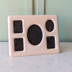 May include: White ceramic picture frame with five openings for photos. The frame has a rounded opening in the center and four rectangular openings around it. The frame has the text "Enchanting Dance" printed on the bottom.