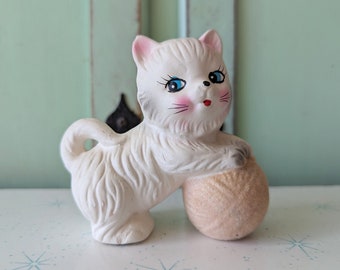 May include: A white ceramic cat figurine with blue eyes and pink cheeks, holding a tan yarn ball. The cat is standing on a light blue surface with a star pattern.