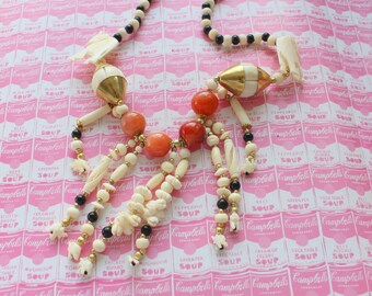 May include: A multi-strand necklace with white and black beads, gold accents, and coral-colored beads. The necklace is photographed on a pink and white background with a repeating pattern of Campbell's Soup labels.