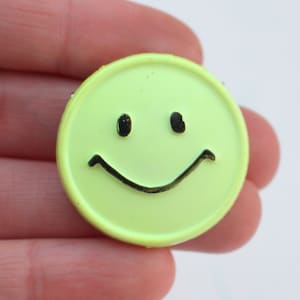 May include: A bright yellow smiley face button with a black outline. The button has a simple design with two eyes and a curved smile.