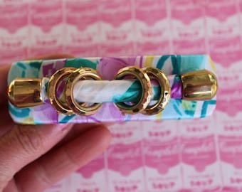 May include: A colorful fabric covered hair clip with gold metal rings and end caps. The fabric has a floral pattern in shades of pink, purple, blue, and green.