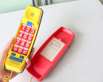 Könnte beinhalten: Ein gelbes Spielzeugtelefon mit einem roten Tastenfeld und einem Spiralkabel. Das Telefon hat einen blauen Knopf und einen Lautsprecher oben. Eine rote Telefonbasis aus Kunststoff ist ebenfalls zu sehen.