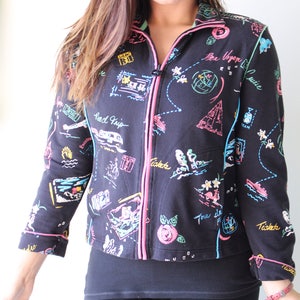 May include: Black zip-up jacket with a colorful travel-themed print. The print features drawings of airplanes, maps, and the words "Road Trip" and "Tickets".