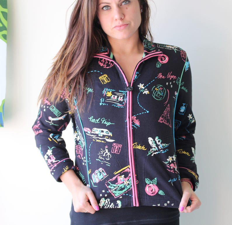 May include: Black zip-up jacket with a colorful travel-themed print. The print features images of airplanes, cars, luggage, and the text "Road Trip" and "Bon Voyage".