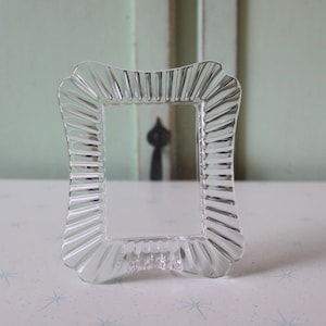 May include: A clear, rectangular picture frame with a scalloped edge design. The frame is made of glass and has a ribbed texture around the border. The frame is approximately 5 inches tall and 3 inches wide.