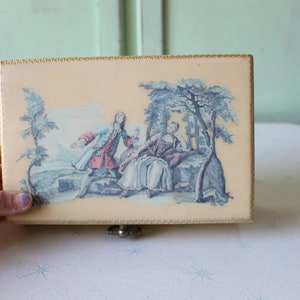 May include: A vintage wooden box with a hinged lid. The lid features a colorful illustration of a couple in a garden setting. The box has a gold trim around the edges.