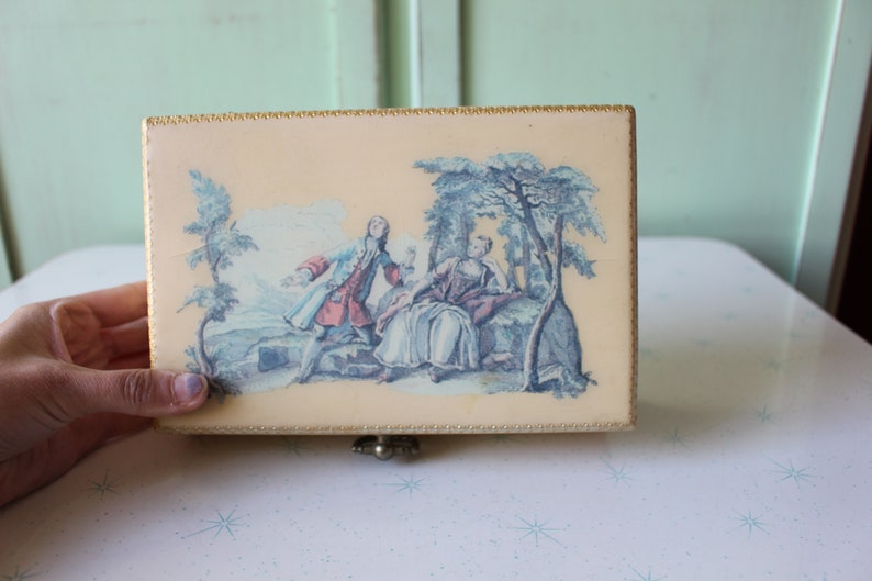 May include: A vintage wooden box with a hinged lid. The lid features a colorful illustration of a couple in a garden setting. The box has a gold trim around the edges.