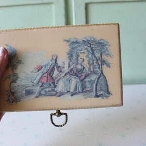 May include: A vintage wooden box with a cream-colored lid featuring a painted scene of two people in a garden setting. The lid has a gold trim and a brass handle.