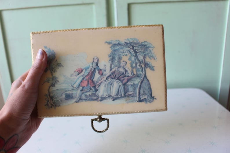 May include: A vintage wooden box with a cream-colored lid featuring a painted scene of two people in a garden setting. The lid has a gold trim and a brass handle.