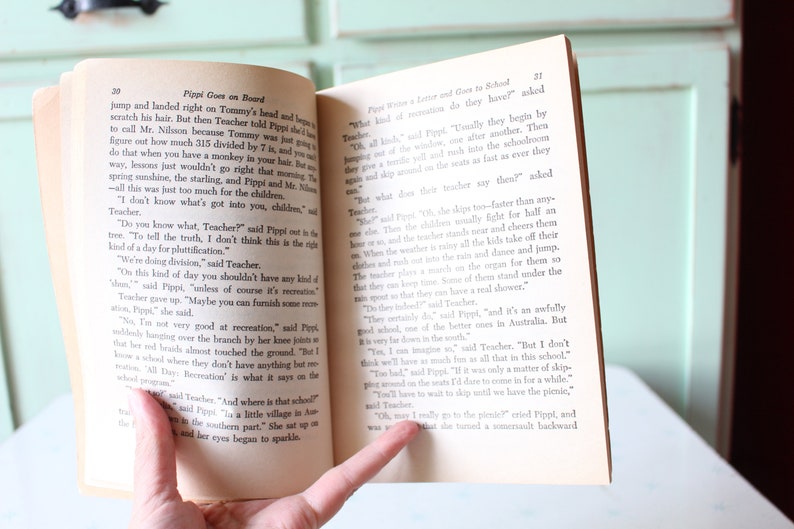 May include: Open book with a yellowed page showing a close-up of a hand holding the book open. The text on the page reads "Pippi Goes on Board" and "Pippi Writes a Letter and Goes to School".