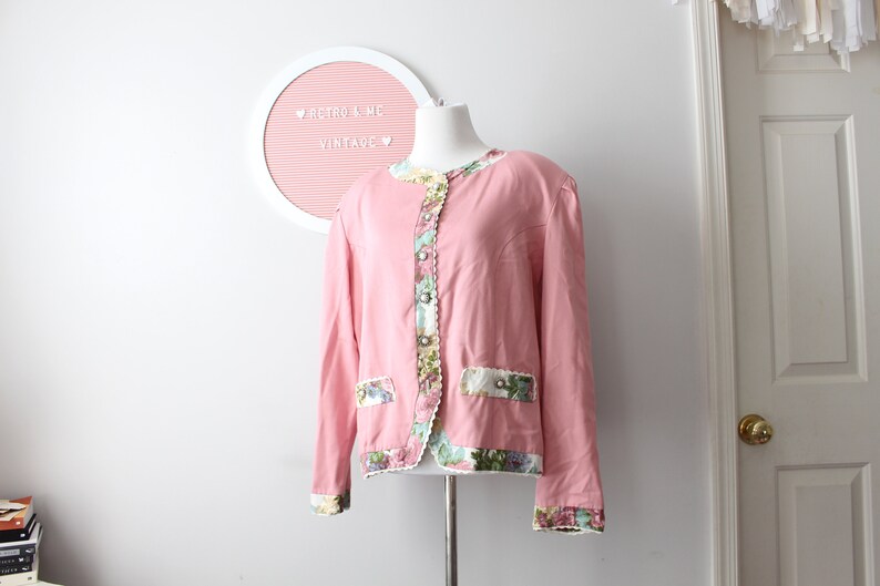 May include: A pink vintage jacket with floral trim and a white lace detail. The jacket has a button closure and long sleeves.