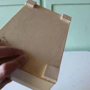 May include: A white rectangular box with four small wooden feet on the bottom. The box is made of a smooth material and has a slightly textured surface.