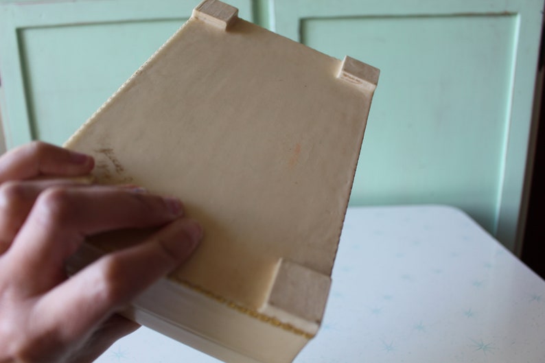 May include: A white rectangular box with four small wooden feet on the bottom. The box is made of a smooth material and has a slightly textured surface.