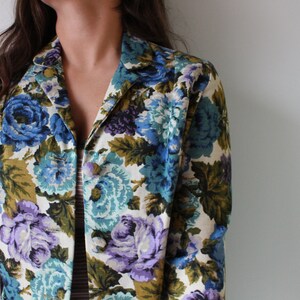 Puede incluir: Una chaqueta vintage de estampado floral con fondo blanco y flores azules, moradas y verdes. La chaqueta tiene cuello y mangas largas.