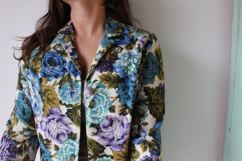 Puede incluir: Una chaqueta vintage de estampado floral con fondo blanco y flores azules, moradas y verdes. La chaqueta tiene cuello y mangas largas.