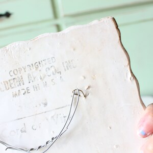 May include: White ceramic wall plaque with a scalloped edge. The back of the plaque has a wire hanger and the text "COPYRIGHTED MODERN ART CO., INC. MADE IN U.S.A."