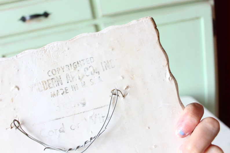 May include: White ceramic wall plaque with a scalloped edge. The back of the plaque has a wire hanger and the text "COPYRIGHTED MODERN ART CO., INC. MADE IN U.S.A."