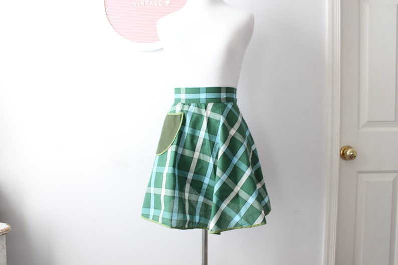 Pode incluir: Uma saia xadrez verde e azul com um bolso verde e uma bainha verde lim&atilde;o. A saia est&aacute; em um manequim branco.