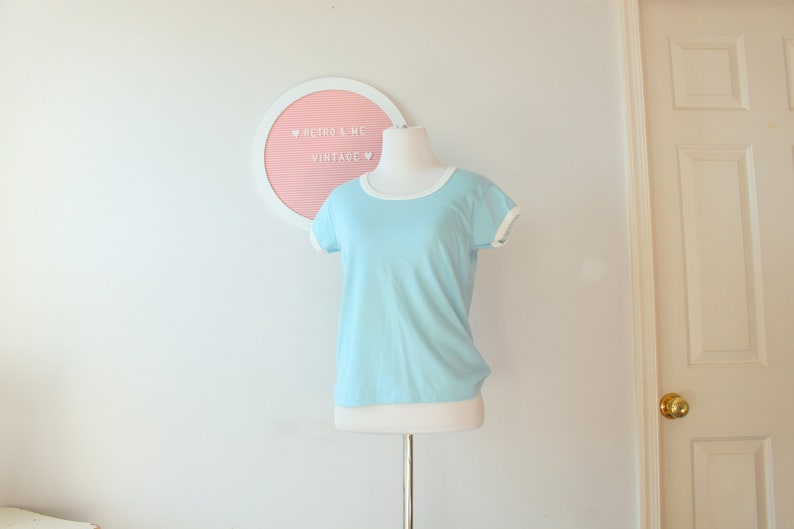 May include: A light blue t-shirt with white trim around the neckline and sleeves. The shirt is on a white mannequin against a white background. The text "Retro & Me Vintage" is on a pink letter board behind the mannequin.