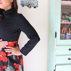 Peut inclure: Une robe noire &agrave; manches longues avec un col montant et un motif floral rouge et noir sur la jupe. La robe a une silhouette ajust&eacute;e. Un meuble de style vintage est en arri&egrave;re-plan.
