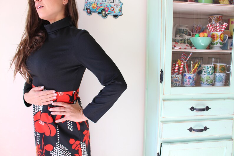 Peut inclure: Une robe noire &agrave; manches longues avec un col montant et un motif floral rouge et noir sur la jupe. La robe a une silhouette ajust&eacute;e. Un meuble de style vintage est en arri&egrave;re-plan.